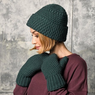 A person wearing the Hand Knit Hush Hat in dark green lambswool with a folded cuff and matching mittens, looking downward. They have short light brown hair and are dressed in a maroon sweater. The background is a textured gray wall.