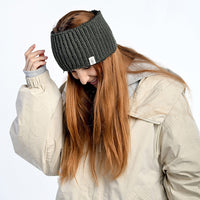 A person with long red hair wears the Hand Loom Mandy Headband, a dark wool accessory handmade in Nepal, along with a light beige winter jacket. They look down with a slight smile against a plain white background.