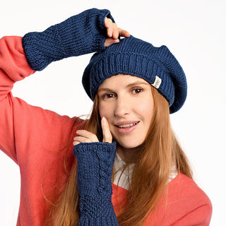A woman with long light brown hair wears a blue knit hat, cozy Cable Handwarmers, and a coral sweater over a white top. She smiles and poses with her hands near her face against a plain white background.