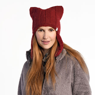 A woman with long auburn hair wears the Ezra Hat—a handmade red wool hat from Nepal featuring ear flaps and cat ear–like pointed corners—paired with a gray fuzzy jacket. She faces forward, smiling slightly against a plain background.