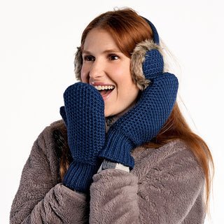 A person with long red hair wears furry earmuffs, a gray fuzzy jacket, and handmade navy blue Kent Mittens, smiling and holding their hands up to their face against a plain white background.
