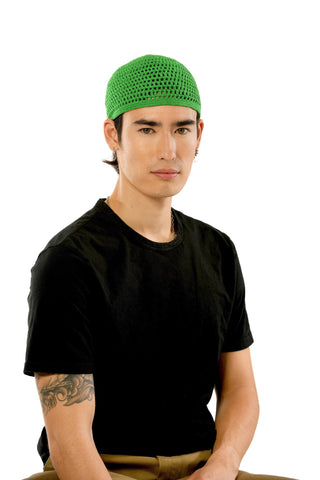 A person with a serious expression is wearing a black T-shirt, a green hand-crocheted Male Beanie, and brown pants. They have a tattoo on their left forearm and are seated against a plain white background.