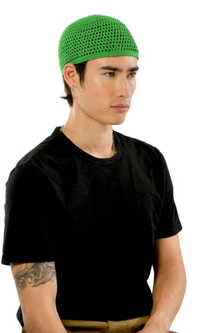 A person with a focused expression is wearing a green Male Beanie from the summer collection and a black t-shirt. They have a tattoo on their left forearm and are sitting against a plain white background.