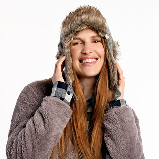 A woman with long red hair smiles while wearing the Fern Hat, a wool hat with ear flaps, paired with a fuzzy gray jacket and plaid shirt. She holds both sides of the hat; the background is white.