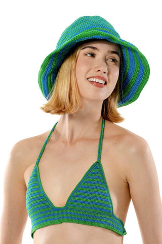 A person wearing a green and blue crocheted halter top and a matching Stripped Sun Hat stands against a white background. The person has medium-length blonde hair, is smiling, and has both arms resting by their sides. They are also wearing denim pants.