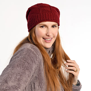 A woman with long red hair wears the Wave Slouch red beanie and a gray fuzzy jacket, smiling at the camera against a white background while holding a section of her hair.