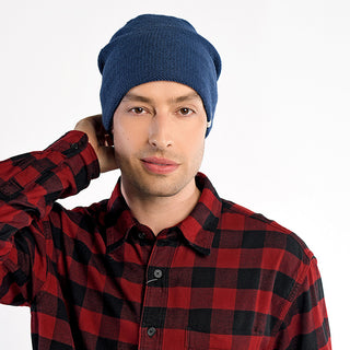 A man wears The Depp Slouch, a blue beanie, and a red and black plaid shirt, standing against a plain white background with one hand behind his neck.