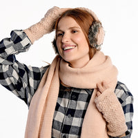 Wearing a black and white plaid shirt, a pale pink knitted scarf, matching handmade fingerless gloves, and Hand Knit Cable Adjustable Earmuffs With Faux Fur in gray, a person smiles and touches their head against a plain white background.