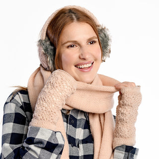 A person wearing Flower Crochet Handwarmers, a pink knit scarf, and fuzzy earmuffs smiles while holding the scarf. They’re also dressed in a black and white plaid shirt against a plain white background.