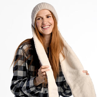 A woman with long red hair smiles while wearing the Oversized Ear Beanie, which features an attached cream-colored scarf. She pairs it with a black and white plaid shirt, standing against a plain white background.