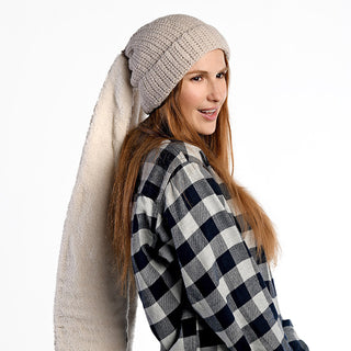 A woman with long brown hair models the Oversized Ear Beanie in beige, featuring an extended tail. She pairs it with a black-and-white checkered shirt and stands against a plain white background, facing slightly to the side.