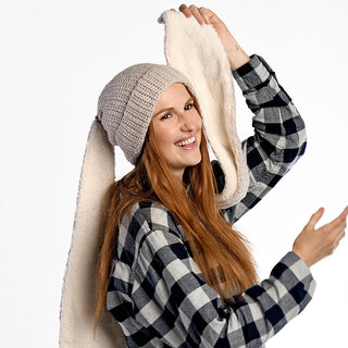 A woman in a plaid shirt wears the Oversized Ear Beanie, featuring long scarf-like extensions, as she holds up one end and smiles at the camera against a plain white background.
