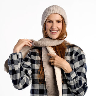 A person with long reddish hair smiles against a white background while adjusting an Oversized Ear Beanie and scarf, paired with a black-and-white plaid shirt.
