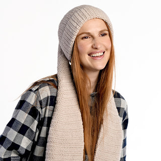 A woman with long red hair wears the Oversized Ear Beanie, featuring cozy hand-knit scarf ends, paired with a black-and-white plaid shirt. She smiles and looks slightly to the side against a plain white background.