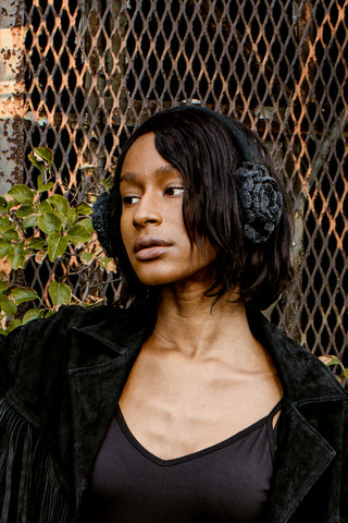 A person with shoulder-length dark hair wears Hand Knit Camellia Earmuffs and a black jacket, standing in front of a rusted metal fence with green leaves visible on the left.