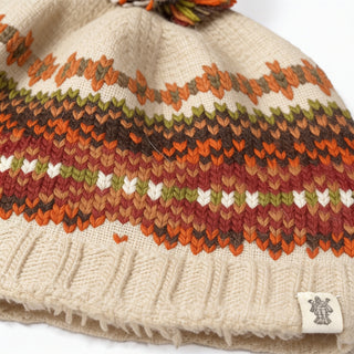 A close-up of the Hand Knit Explore Hat in beige, showcasing a colorful zigzag pattern in orange, brown, green, and white. A small fabric tag with a logo is sewn onto the ribbed edge at the bottom.