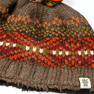 A close-up of the Hand Knit Explore Hat, a brown Fair Isle beanie adorned with orange, green, and white patterns. This hand knit wool hat showcases a small white fabric tag bearing a logo and is finished with a pom-pom crafted from coordinating yarn colors.