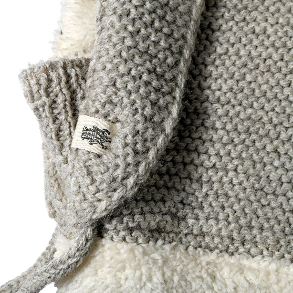 Close-up of the Hand Knit Glide Hood, showcasing its gray wool seed-stitch pattern with a prominent white label featuring a black tree motif. The hand-knit texture is evident, along with some fluffy white material visible underneath.