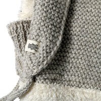 Close-up of the Hand Knit Glide Hood, showcasing its gray wool seed-stitch pattern with a prominent white label featuring a black tree motif. The hand-knit texture is evident, along with some fluffy white material visible underneath.