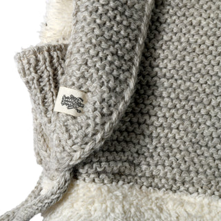 Close-up of the Hand Knit Glide Hood, showcasing its gray wool seed-stitch pattern with a prominent white label featuring a black tree motif. The hand-knit texture is evident, along with some fluffy white material visible underneath.