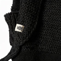 Close-up of the Hand Knit Glide Hood in black, showcasing its textured hand knit fabric and a visible white tag with a black graphic design. The details of the knit and fabric edges are clearly displayed against a white background.
