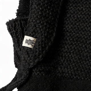Close-up of the Hand Knit Glide Hood in black, showcasing its textured hand knit fabric and a visible white tag with a black graphic design. The details of the knit and fabric edges are clearly displayed against a white background.