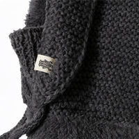 Close-up of the Hand Knit Glide Hood in dark gray, showcasing the chunky seed-stitch texture and a small white fabric tag with a black illustration attached to the garment. The hand-knit craftsmanship is clearly visible.