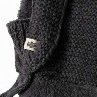 Close-up of the Hand Knit Glide Hood in dark gray, showcasing the chunky seed-stitch texture and a small white fabric tag with a black illustration attached to the garment. The hand-knit craftsmanship is clearly visible.
