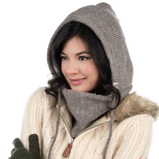 A woman wearing a gray Hand Loom Hero Hood, a matching scarf, a cream sweater with a Sherpa fleece-lined hood, and green gloves looks sideways with a neutral expression against a white background.