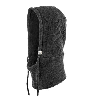 The Hand Loom Hero Hood is a dark gray, knitted Merino wool hood with a drawstring and front tag, shown on a white background. Its thick, textured fabric offers extra warmth for winter.