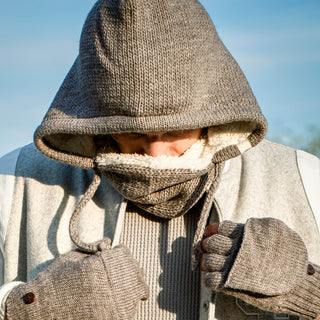 Someone bundled up against the cold wears the Hand Loom Hero Hood, a scarf, and fingerless gloves outdoors. Their face is mostly covered, looking down with a blue sky in the background.