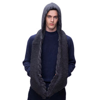 Wearing a navy blue sweater and the Hand Knit 2 Pocket Scarf Hood—a chunky, dark gray merino wool scarf with a hood, sherpa lining, and handmade in Nepal—the person stands against a white background and looks directly at the camera.