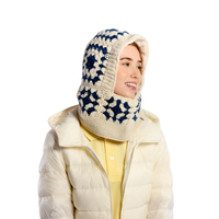 A person wearing the Hand Crochet Patch Hood—a cream and navy blue hand-crocheted wool hooded scarf—paired with a cream puffer jacket over a yellow collared shirt, facing slightly to the right and smiling against a plain, light-colored background.