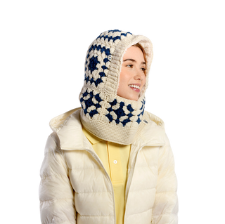 A person wearing a Crochet Patch Hood, a white knitted garment with blue geometric patterns and a hood. They are also wearing a white padded jacket over a yellow shirt against a black background.