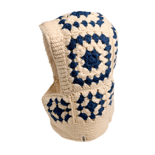 The Crochet Patch Hood showcases a cream base adorned with blue diamond patterns. This crocheted balaclava design incorporates yarn-crafted squares and has an opening at the face, set against a plain backdrop.