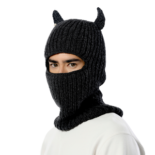 A person is wearing the Hood With Horns, a handcrafted wool balaclava with small pointed horns on top. The balaclava covers their head, neck, and lower face, leaving only the eyes exposed. They are dressed in a white top and are positioned against a black background.