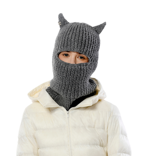 A person is wearing the Hood With Horns, a gray knit balaclava featuring pointy ears, along with a white puffer jacket—together making an intriguing winter accessory. Only the eyes and part of the forehead are visible against a black background.