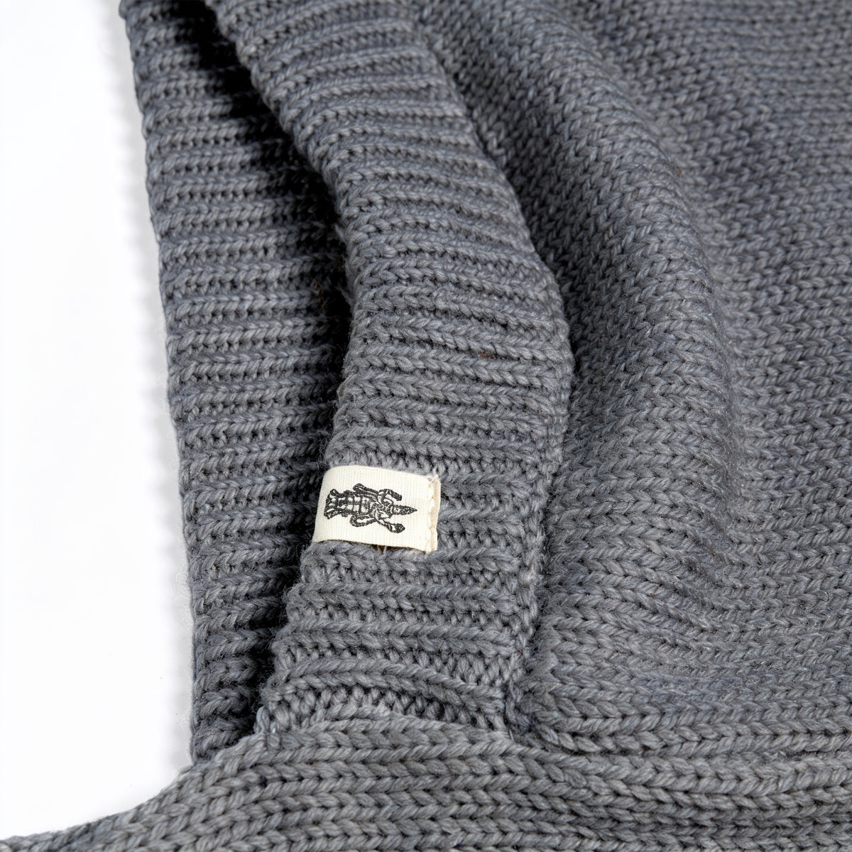 A close-up of the gray knitted fabric of the Hand Loom Nomad Hood Scarf, with a small white tag featuring a black fish illustration attached to the edge. The fabric appears folded, showcasing the detailed texture of the knit.