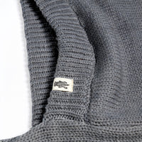 A close-up of the gray knitted fabric of the Hand Loom Nomad Hood Scarf, with a small white tag featuring a black fish illustration attached to the edge. The fabric appears folded, showcasing the detailed texture of the knit.