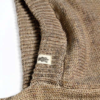 A close-up of the Hand Loom Nomad Hood Scarf, a knitted beige piece with a small rectangular tag sewn onto the ribbed edge. The tag displays a black graphic of a crab, and the visible knit pattern and fibers highlight craftsmanship reminiscent of hand knitting.