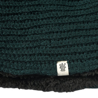 A close-up of the Hand Knit Halo Reverse Hood reveals dark green knitted fabric with a small white tag displaying a simple black graphic. The bottom edge features black textured material, possibly fleece or faux fur, highlighting the cozy finish of this hand-knit hood.