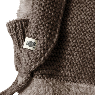 Close-up of the Hand Knit Glide Hood, a brown hand-knit garment featuring a textured seed-stitch pattern, woolly lining, and a small white fabric tag with a black embroidered logo stitched into the seam.
