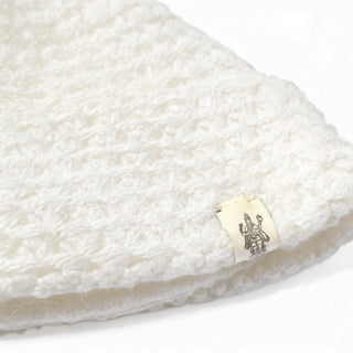 Close-up of a white, crocheted fabric item with a small, off-white tag featuring a printed grey logo. The tag is sewn into the edge of the fabric, reminiscent of detailing found on the Hand Knit Hush Hat—a lambswool beanie with a neatly folded cuff. The background is plain white.