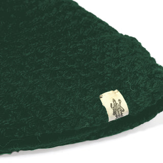 A close-up of the Hand Knit Hush Hat in dark green, showcasing its hand knit texture and a small beige tag with an intricate dark design, placed at the edge of the folded cuff.