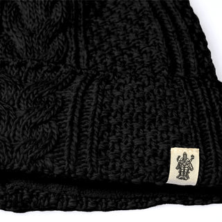 Close-up of the Hand Knit Inter hat in black, showcasing a distinct cable knit pattern. A small beige tag adorned with an intricate illustration of a multi-armed figure is sewn onto the edge, emphasizing its artisanal craftsmanship. This hand-knit wool beanie offers both stylish design and cozy warmth—ideal for cooler days.