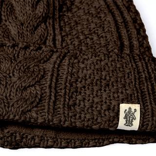 A close-up of the Hand Knit Inter hat, showcasing its brown wool construction with a textured pattern and a small white tag featuring a black line drawing of a figure with multiple arms. The edges of the fabric are clearly visible.