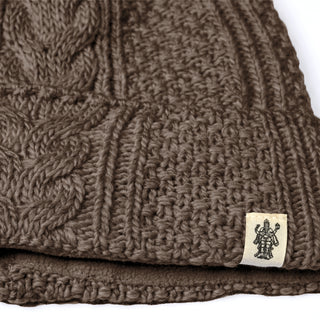 Close-up of the Hand Knit Inter hat, a brown cable knit beanie showcasing a braided cable pattern and finished with a small beige tag adorned with a black printed illustration of a multi-armed figure. The thick, textured hand-knit wool fabric suggests this winter hat delivers both warmth and style.