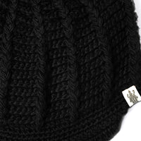 Close-up of the Hand Crochet Martha Cap in black, featuring vertical ribbed knitted fabric reminiscent of a handmade wool hat. A small white tag with a printed illustration of a person holding an object is attached to the edge. The background is white.