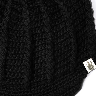 Close-up of the Hand Crochet Martha Cap in black, featuring vertical ribbed knitted fabric reminiscent of a handmade wool hat. A small white tag with a printed illustration of a person holding an object is attached to the edge. The background is white.