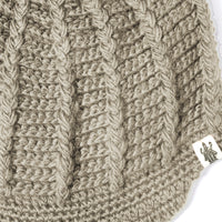Close-up of the Hand Crochet Martha Cap in beige, highlighting its intricate braided knit pattern and a small white tag on the bottom right with an illustration of a person holding an object—an ideal display of the exceptional craftsmanship that defines this handmade wool hat.